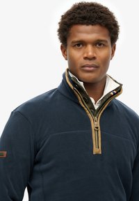 Mørkeblå fleecepullover med kvart lynlås, brunt halsudskæringsdetalje og grøn foring. Har en lynlås og et lille logo på venstre ærme.
