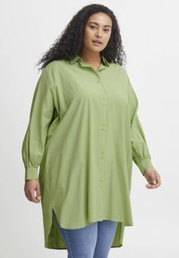 Camicia verde con bottoni, maniche lunghe con polsini bottonati, vestibilità ampia, fessure laterali e trama liscia, abbinata a jeans blu.