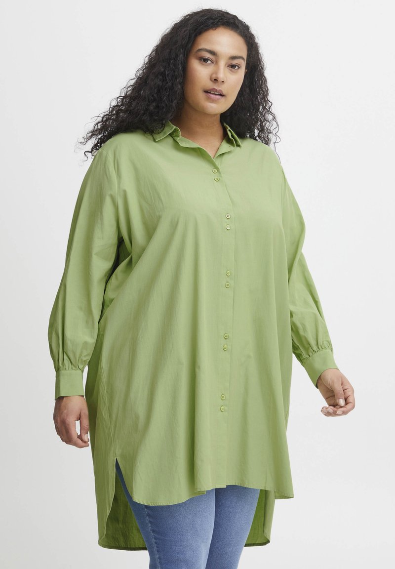 Camicia verde con bottoni, maniche lunghe con polsini bottonati, vestibilità ampia, fessure laterali e trama liscia, abbinata a jeans blu.