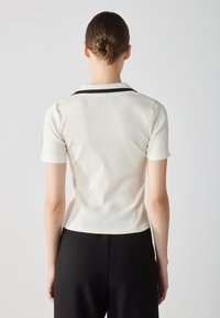 White ribbed short-sleeve polo shirt with a black collar detail, fitted design, and smooth texture, worn with black trousers, view from the back.