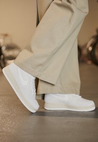 Białe zamszowe sneakersy o teksturowanym wykończeniu, z kremową gumową podeszwą i białym logo Nike, zestawione z szerokimi beżowymi spodniami.