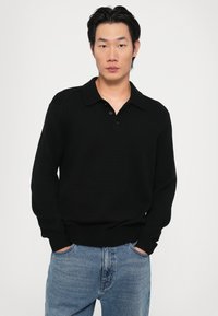 Pulover negru tricotat cu guler și deschidere cu trei nasturi, asortat cu blugi denim albaștri. Textura este dublată și ajustată.