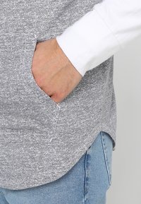 Grå texturerad hoodie med en vit ärm, som har en sidficka och en avslappnad passform. Tyget har en subtilt mönstrad design.