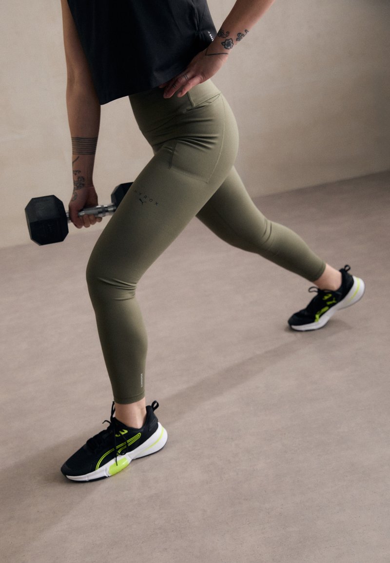 Oliven grønne leggings med sidelommer, kombinert med svarte og neongrønne treningssko. Figuren holder en svart dumbbell.