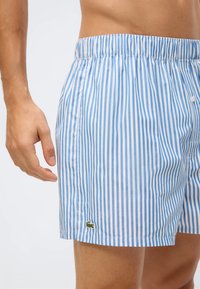 Μπλε και λευκές ριγέ μαγιό κοντό παντελόνι με ελαστική μέση, που διαθέτει ένα μικρό πράσινο λογότυπο στο κάτω άκρο. Απαλό βαμβακερό υλικό.
