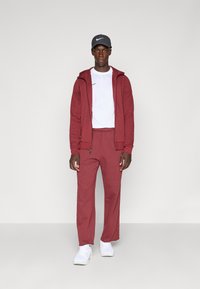 Bordeauxfarbener Reißverschluss-Hoodie und passende Jogginghose aus einem weichen, strukturierten Stoff; getragen über einem weißen T-Shirt und kombiniert mit weißen Sportschuhen und einer grauen Mütze.