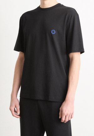 Mann trägt ein einfaches schwarzes T-Shirt mit kurzen Ärmeln und kleinem blauem Smiley-Logo auf der linken Brust, steht vor weißem Hintergrund.