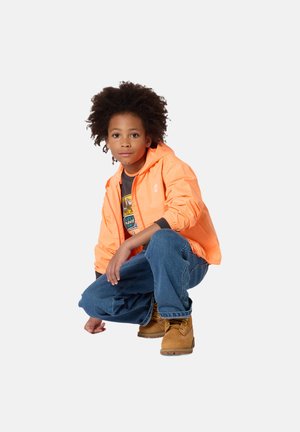 Enfant aux cheveux bouclés accroupi, portant une veste orange, un jean bleu, des bottes beige et un t-shirt à motif, sur un fond clair uni.