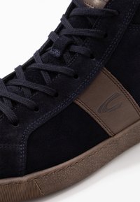 Námořnické semišové tenisky s hnědým koženým bočním panelem, texturovanou gumovou podrážkou a kontrastním prošíváním. Šňůrky a jazyk jsou také námořnické barvy.