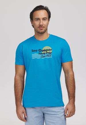 Mężczyzna noszący jaskrawoniebieską koszulkę "Lee Cooper Sunny Days Since 1908" i jasnoniebieskie spodnie, stojący na tle jednolitego szarego tła.