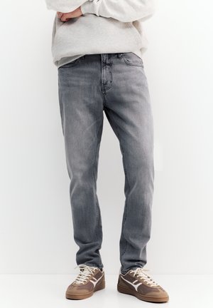 Egy férfi szürke farmert, világosszürke pulóvert és barna sportcipőt visel, és keresztbe tett karokkal áll egy egyszerű fehér háttér előtt.