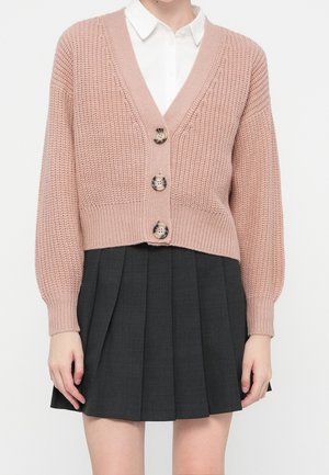Femme portant un gilet en maille épaisse rose clair avec de gros boutons, par-dessus une chemise blanche à col et une jupe plissée gris foncé.