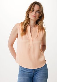 Brzoskwiniowa, bezrękawnikowa bluzka z kołnierzykiem mandarynkowym, z pionowym dekoltem oraz gładką fakturą, zestawiona z jasnoniebieskimi dżinsami.