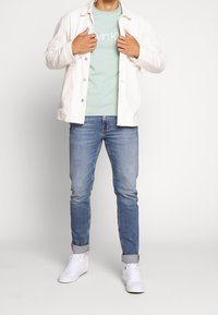 Chaqueta de algodón blanca sobre una camiseta de color verde claro con logo, combinada con jeans de mezclilla azul claro y zapatillas blancas. Mangas cortas, enrolladas.