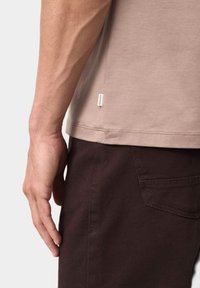 Beżowa koszulka z krótkim rękawem o delikatnej teksturze, zestawiona z ciemno-brązowymi spodniami. Zawiera małą metkę marki przy dolnym brzegu.