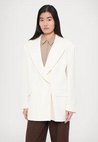 Biely dvojradový blejzer s výrezmi na klopách, bočnými vreckami a štruktúrovaným strihom, vrstvený cez hnedastú košeľu a hnedé nohavice.