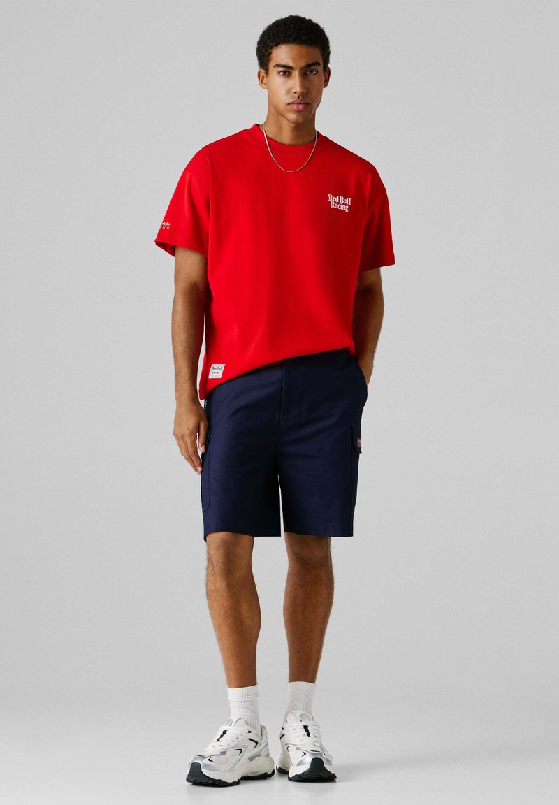 Homme portant un T-shirt rouge Red Bull Racing, un short bleu marine, des chaussettes blanches et des chaussures de sport blanches, debout devant un fond gris uni.