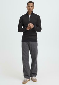 Čierny sveter na zips z textúrovaného materiálu, kombinovaný so sivými nohavicami s širokými nohavicami. Oblečenie dopĺňajú hnedé tenisky. Jednoduchý, moderný dizajn.
