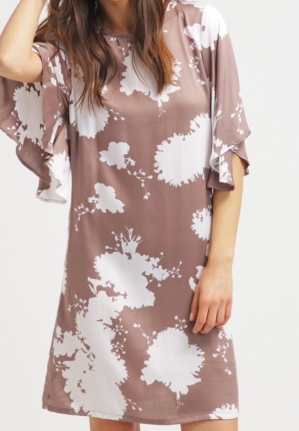 Femme portant une robe mauve mi-longue avec un grand imprimé floral blanc et des manches flottantes, debout devant un fond clair uni.