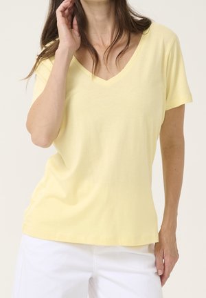 Mujer con una camiseta de manga corta, de color amarillo claro con cuello en V y pantalones blancos, con una mano tocándose el pelo y la otra a un lado.