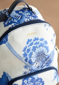 Mochila con estampado floral azul sobre un fondo blanco, que cuenta con un compartimento principal con cremallera, asas de tela y un detalle del logotipo en dorado.