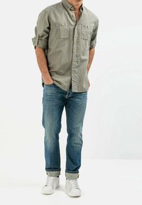 Lichtgrijs overhemd met lange mouwen en knopen, voorzien van opgerolde mouwen en twee borstzakken. Draagt blauwe spijkerbroek en witte sneakers.