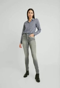 Camisa de botão em xadrez azul-marinho e branco, combinada com jeans skinny cinzentos de alta cintura e fecho de cinco botões. Botins pretos.