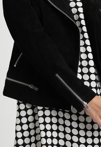 Svart mocka bikerjacka med silverfärgade dragkedjor, med en skräddarsydd design och texturerad yta. Underjackan syns en vit klänning med svarta prickar.