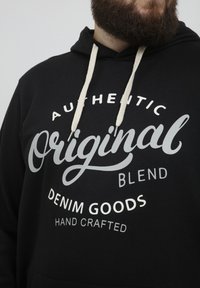 Sudadera negra con cordones blancos y un destacado texto gráfico plateado: "Auténtico Original Blend Denim Goods Hecho a Mano."