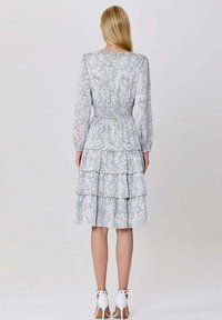 Femme aux longs cheveux blonds portant une robe longue à manches longues bleu clair à fleurs avec une jupe à volants et des sandales à talons blancs, se tenant de dos.