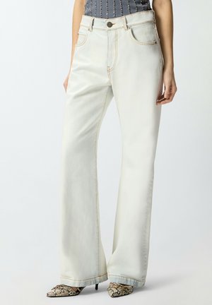 Donna che indossa jeans a vita alta, gamba larga, colore avorio chiaro, e tacchi a punta effetto pelle di serpente, con una maglia scura impreziosita da piccole perline.