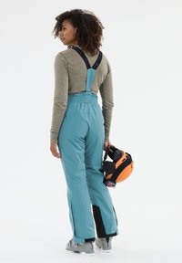 Turkosa skidräkter med svarta remmar, elastisk midja, siddragkedjor och ett mjukt material. Modellen håller en orange hjälm.