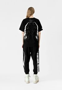 Camiseta negra con gráficos de alas en blanco y negro en la parte posterior, combinada con pantalones negros que tienen detalles de alas y una etiqueta, llevados con botas blancas.