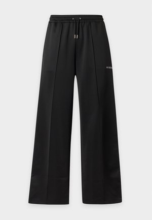 Mann steht mit den Händen in den Taschen, trägt ein weißes kurzärmeliges Hemd, schwarze weite Hosen und schwarze Sneakers mit weißen Sohlen.