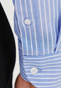 Niebiesko-biała materiał w paski z guzikami na mankiecie. Tekstura wydaje się gładka, a paski są równomiernie rozmieszczone.