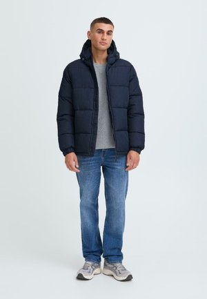 Marineblaue Daunenjacke mit Kapuze, ausgestattet mit gesteppten Segmenten und elastischen Bündchen, getragen über einem grauen Hemd und kombiniert mit blauen Jeans und Sneakers.
