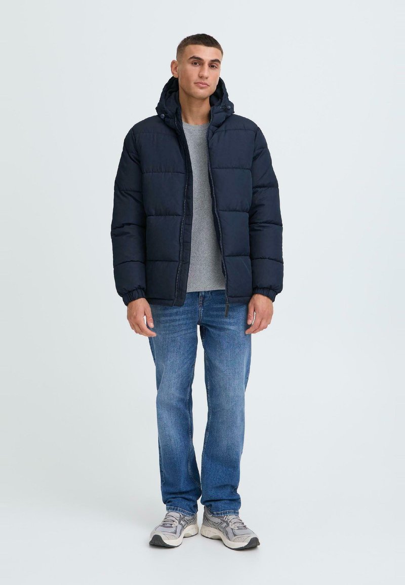Marineblå puffjakke med hætte, præget af kviltede sektioner og elastiske manchetter, båret over en grå skjorte og parret med blå jeans og sneakers.