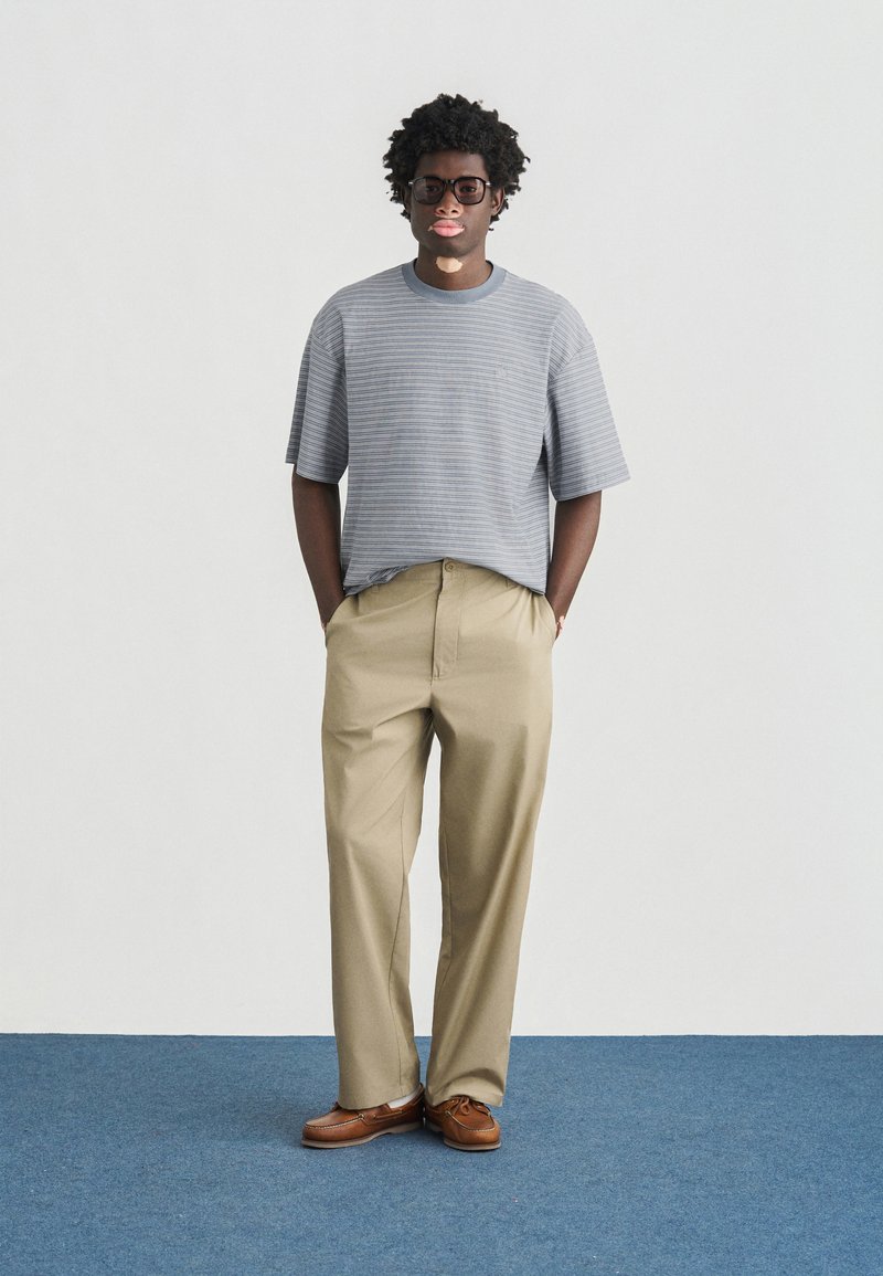 Gray and white striped short-sleeve shirt, beige trousers, brown shoes, dark-framed glasses, hands in pockets, neutral backdrop.