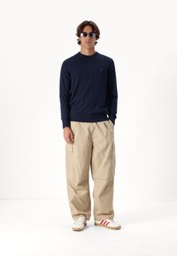 Námořnický svetr, béžové cargo kalhoty a bílé tenisky s červenými pruhy. Svetr má kulatý výstřih a přiléhavé manžety, cargo kalhoty mají boční kapsy.