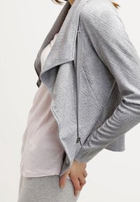 Šedá bunda na zip s texturovanou tkaninou, zavinovacím límcem a přiléhavým střihem, nošená přes světle růžový top.