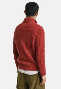 Rode kabelgebreide trui met een hoge kraag en geribbelde manchetten, voorzien van een gestructureerd patroon, gedragen over een wit shirt, met donkere broek.