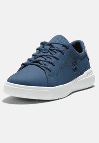 Zapatilla azul con parte superior sintética suave, lengua texturizada, suela de goma blanca y detalle del logotipo en el costado. Presenta cordones tradicionales.