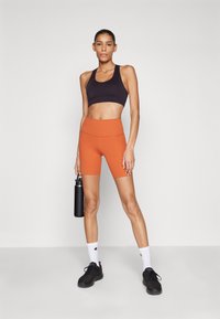 Bra de sport noir, short taille haute orange, baskets noires. Le modèle tient une bouteille d'eau noire, debout sur un fond uni clair.