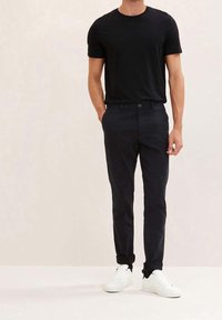 Maglietta nera e pantaloni blu scuro. I pantaloni hanno una tasca frontale; entrambi i capi sono aderenti. Le sneakers bianche aggiungono contrasto ai piedi.