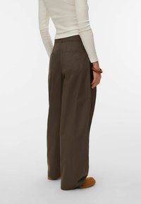 Personne portant un pantalon large marron et un haut blanc à manches longues, debout avec un bras détendu et portant un bracelet en bois.