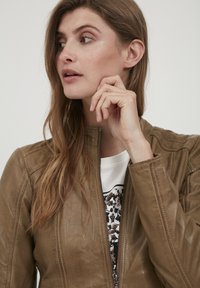 Hnedá kožená bunda so zipsom vpredu, okrúhlym golierom a postrannými švy. Nosená na bielej košeli s dekoratívnym vzorom.