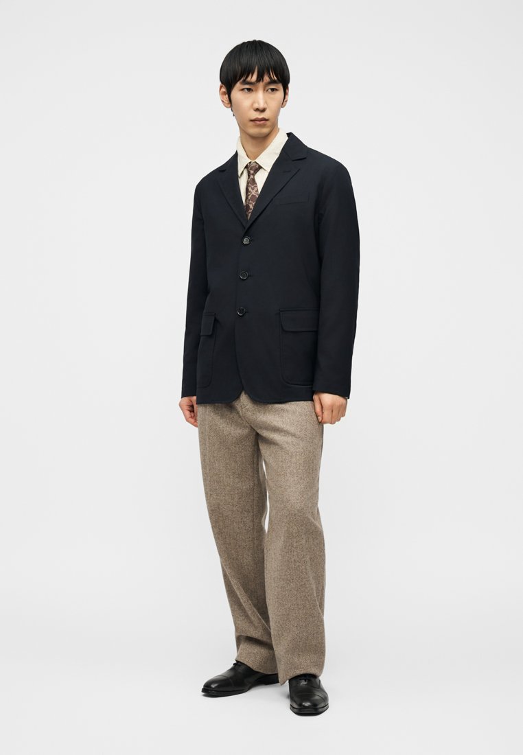 Man wearing black blazer, beige patterned tie, cream shirt, brown tweed trousers, and black leather shoes standing against white background.