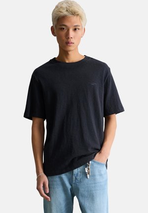 Ung voksen med kort blegnet hår iført en løs sort t-shirt og lyseblå jeans med nøgler fastgjort, stående mod en ensfarvet baggrund.