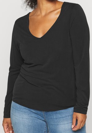 Vrouw draagt een effen zwart, lang mouwig V-hals top en lichtblauwe spijkerbroek, afgebeeld van nek tot heupen tegen een lichte achtergrond.