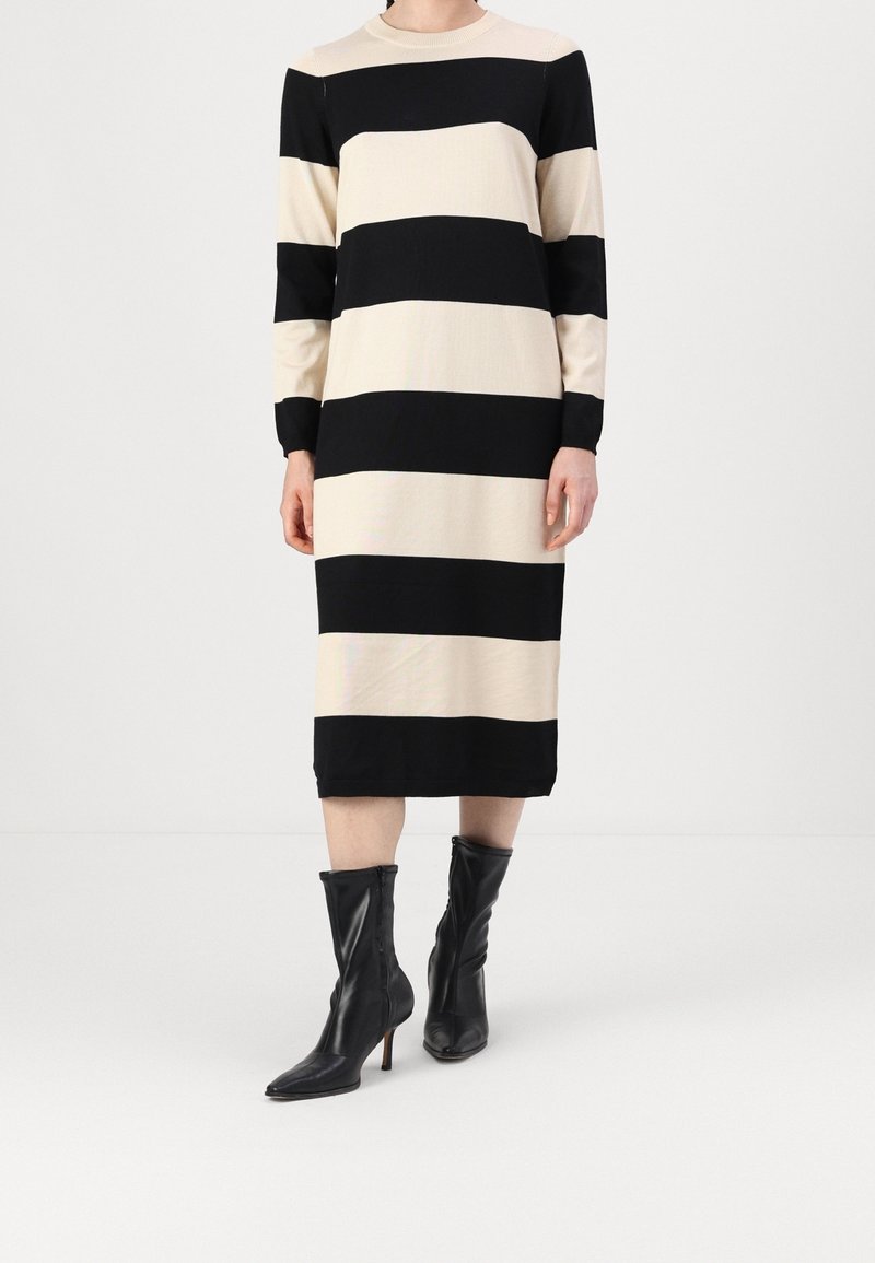 Personne portant une robe à manches longues, longueur genoux, avec de larges rayures horizontales noires et beiges et des bottines noires à bout pointu avec talons.